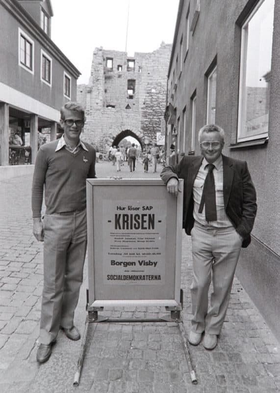 Ingvar Carlsson och Kjell-Olof Feldt står på var sida om en skylt med texten "Hur löser SAP krisen. Borgen Visby. Socialdemokraterna". I bakgrunden syns ett av stadsmurens torn.